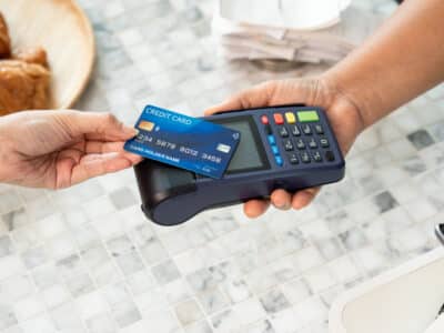 Two adult hands exchanging payment using blue credit card with wireless symbol on electronic POS terminal placed on marble table surface in cafe environment with croissant plate in background